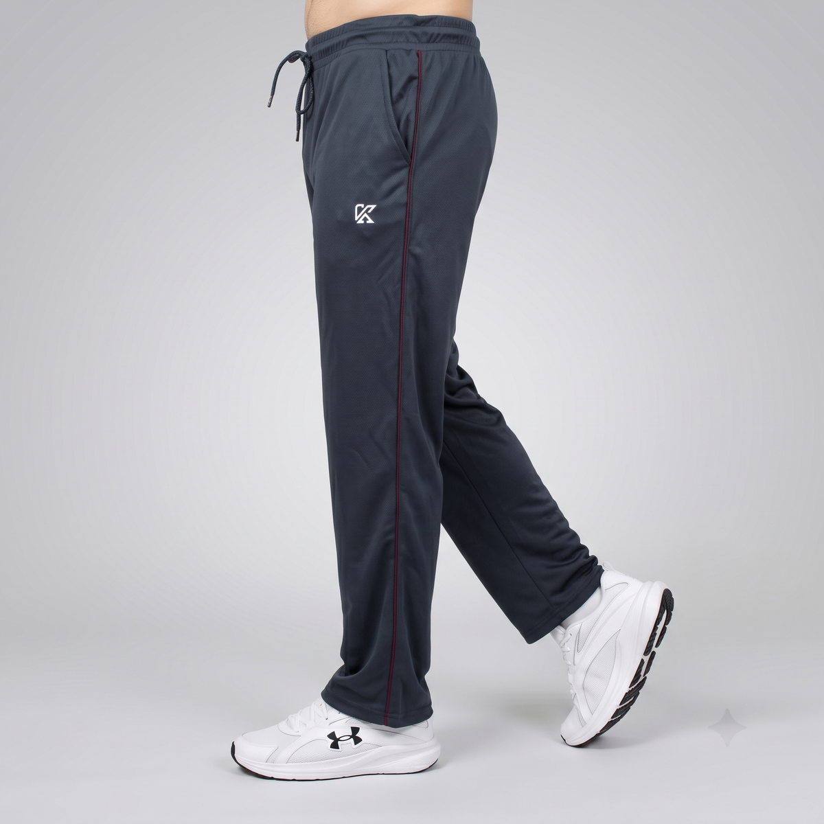 A person shown from the waist down wearing dark slate gray athletic sweatpants with a white embroidered logo on the left thigh and white Under Armour sneakers, captured against a light gray studio background.