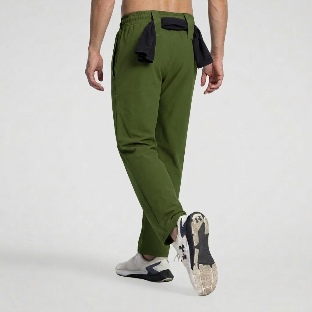 A man wearing olive green athletic jogger pants with black side details and white and navy sneakers, shown from a rear three-quarter angle against a light gray studio background.