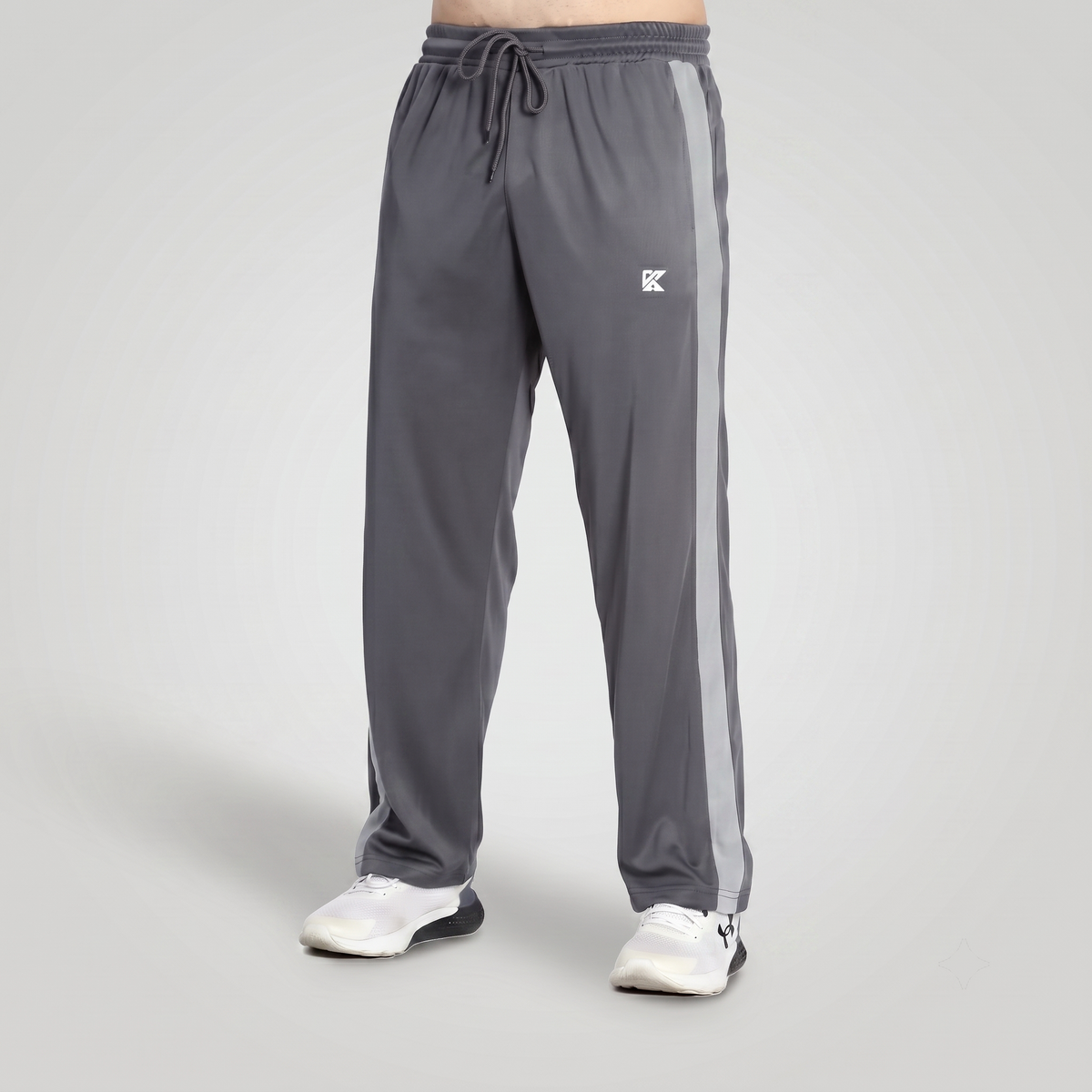 Gray athletic track pants with a white side stripe and a small white logo on the left thigh, shown on a model from the waist down wearing white sneakers against a light gray studio background.