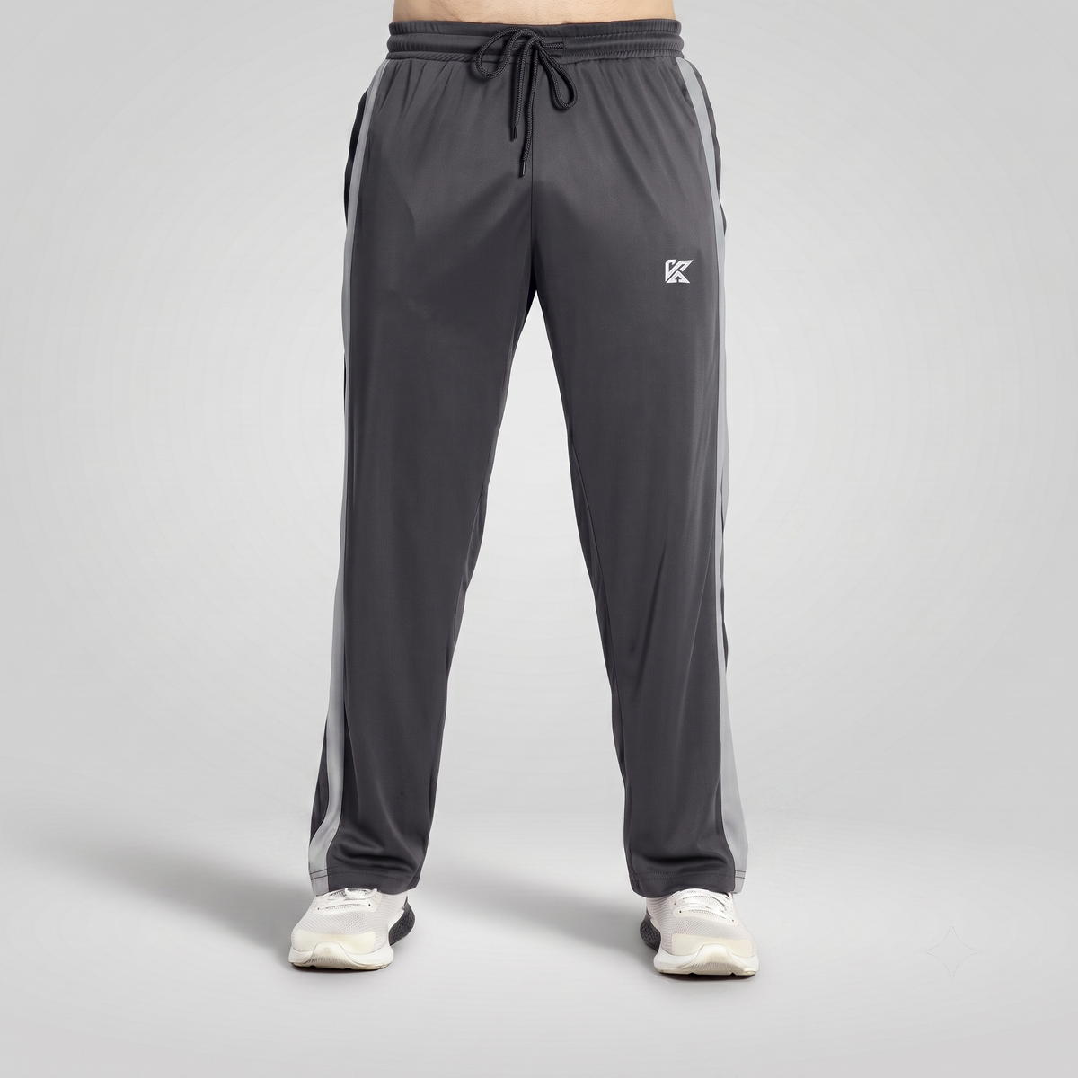 Dark gray athletic sweatpants with white side stripes and a white logo on the left thigh, shown on a model from the waist down wearing white sneakers against a light gray studio background.