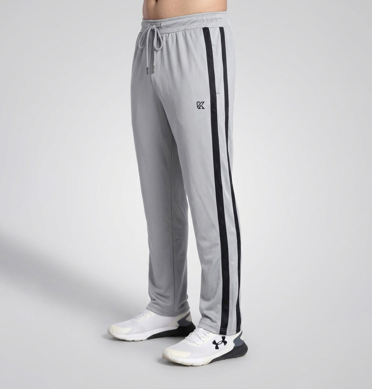 A person wearing light gray athletic sweatpants with two black side stripes and a small black logo on the upper left thigh, paired with white sneakers, photographed against a light gray studio background.