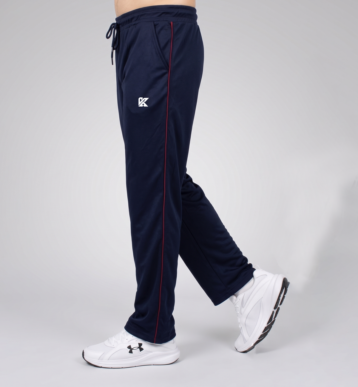 A person wearing navy blue athletic jogger pants with a thin red side stripe, a white embroidered logo on the left thigh, and white sneakers, shown from the waist down against a light gray studio background.